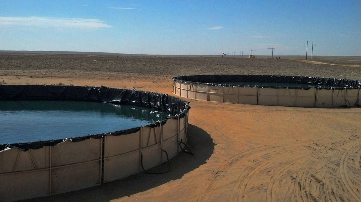 Two filled tanks with black liners on an open desert location under a clear sky, with hose connections at the base.