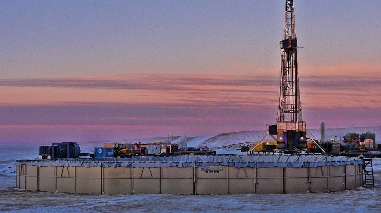 A Hydro Towers modular water storage tank on a snow-covered pad beside an active drilling rig at dusk.