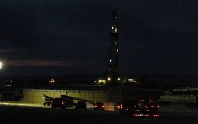 Tanks lit at night beside a drilling rig, with vehicles in the foreground.