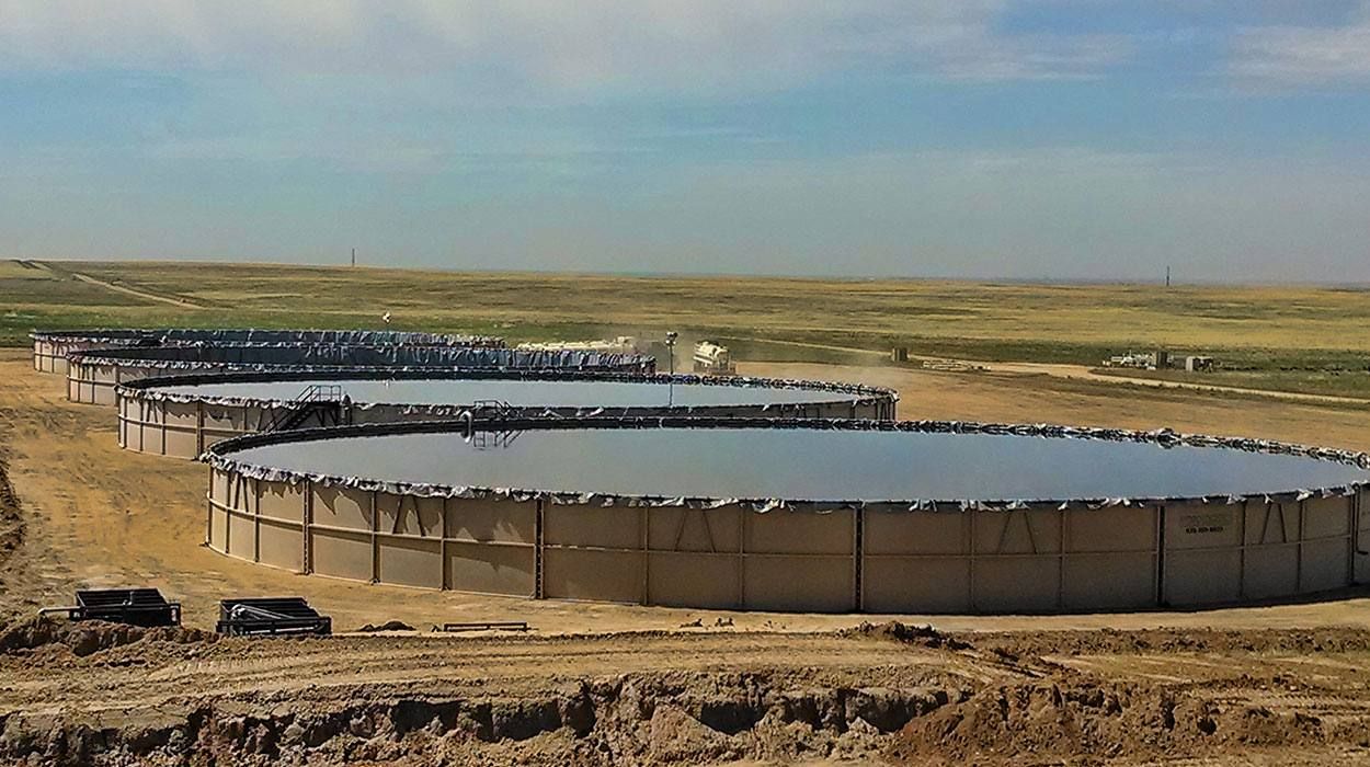 Four modular tanks set in a line on a prepared pad, filled with water, with open grassland beyond.