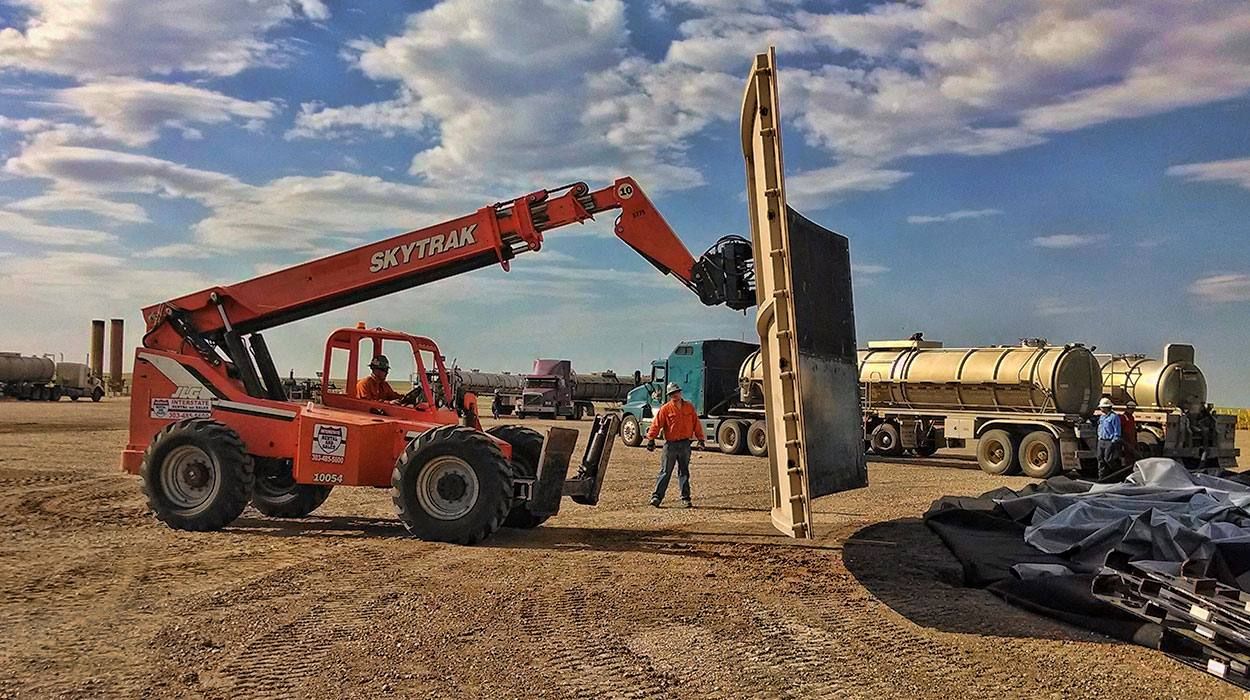 A crew member in high-visibility clothing guides a tank panel as a telehandler lifts it into position, with water trucks waiting behind.